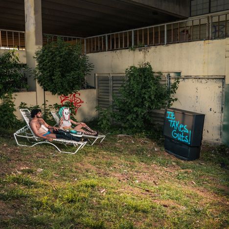 Texte: "It TAKES Guts!" auf einem alten Fernseher. Zwei Personen liegen auf Sonnenliegen in einem verwilderten, urbanen Umfeld.