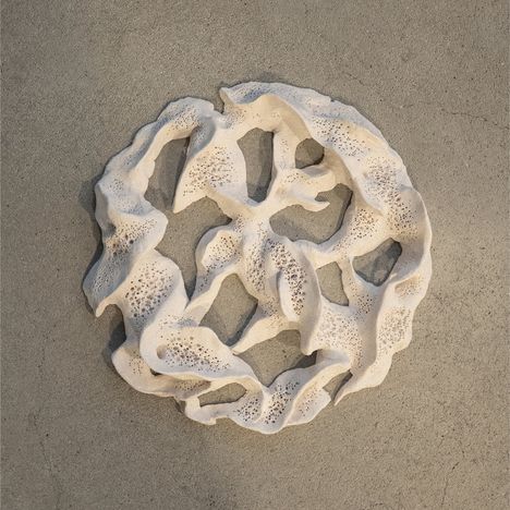 Eine dekorative, cremefarbene, durchbrochene Form liegt auf grauem Hintergrund, erinnert an filigranes Korallenmuster.