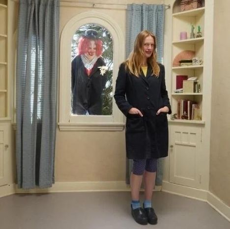 Ein Raum mit Regalen, eine Frau steht vorne, ein Clown schaut durchs Fenster. Beide tragen dunkle Mäntel.