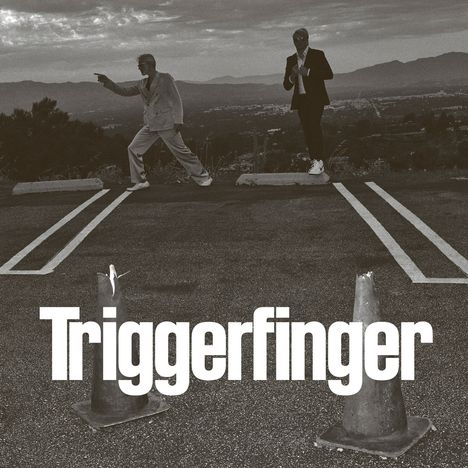 Text "Triggerfinger". Zwei Männer in Anzügen stehen auf einem Parkplatz, einer zeigt mit dem Finger, im Hintergrund Berge.