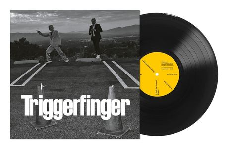 "Triggerfinger" in großen Buchstaben. Zwei Männer im Anzug auf einer Straße, im Hintergrund eine Stadtansicht.