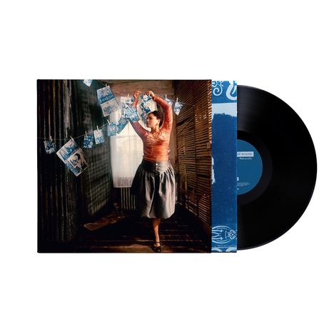 „Naturally“ Album. Frau in Tanzpose hängt Fotos auf. Vinyl-Schallplatte daneben. Vintage-Atmosphäre in einem Raum.