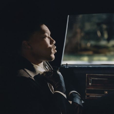 Keine Texte vorhanden. Ein Mann in Anzug und Handschuhen sitzt in einem Auto und blickt nachdenklich aus dem Fenster.