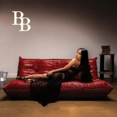Text "BB". Frau in schwarzem Kleid liegt auf rotem Sofa. Bücher stapeln sich auf kleinem Tisch daneben. Schlichte Umgebung.