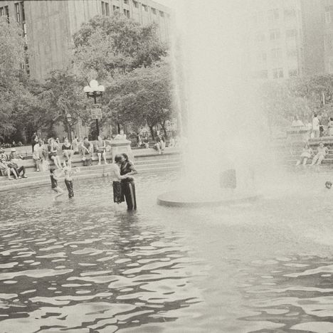 Ein Park mit einem großen Springbrunnen, viele Menschen sitzen und stehen herum, Kinder spielen im Wasser.
