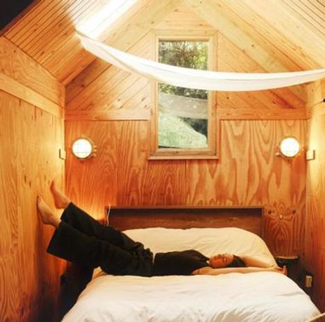 Keine erkennbaren Texte. Eine Person liegt entspannt auf einem Bett in einem holzgetäfelten Raum mit schräger Decke.