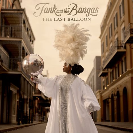 „Tank and the Bangas: The Last Balloon“. Eine Frau mit Federkopfschmuck hält einen silbernen Ballon in einer eleganten Straße.