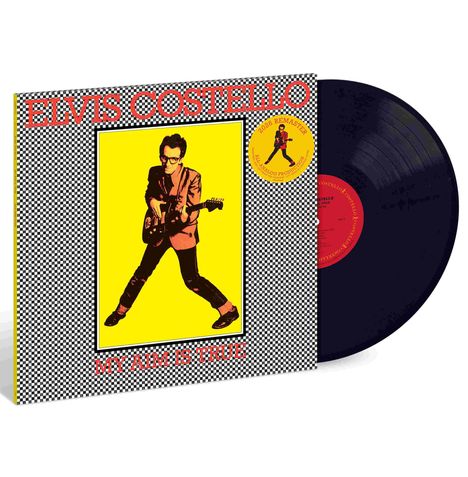 "Elvis Costello" oben, "My Aim Is True" unten. Illustration eines Mannes mit Gitarre auf gelbem Hintergrund. Schallplatte sichtbar.