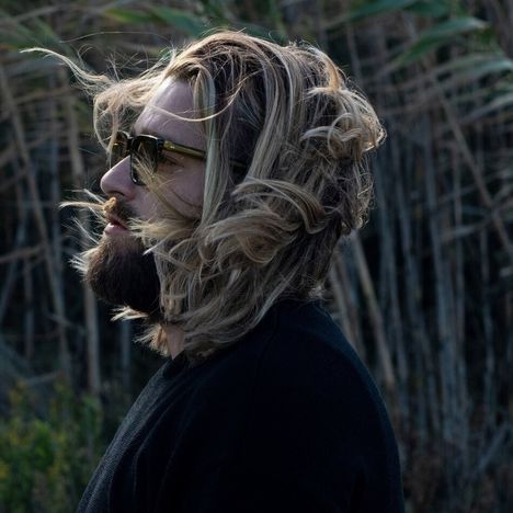 Ein Mann mit langen blonden Haaren und Sonnenbrille blickt seitlich, Hintergrund aus Pflanzen und Schilf.