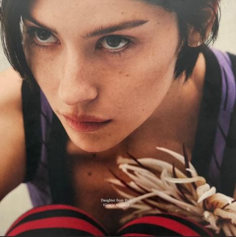 „Daughter from Hell“. Frau mit kurzem, dunklem Haar, intensiver Blick. Sie hält ein weiß-rotes, blumenartiges Objekt.