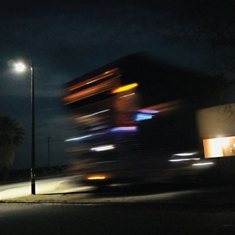 Keine Texte vorhanden. Verschwommener, beleuchteter LKW bei Nacht, vorbei an einer Straßenlaterne und beleuchtetem Gebäude.
