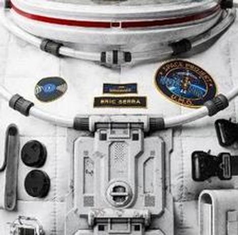 Text: "ERIC SERRA" auf einem Namensschild. Raumanzug mit NASA-Patch und technischen Details.