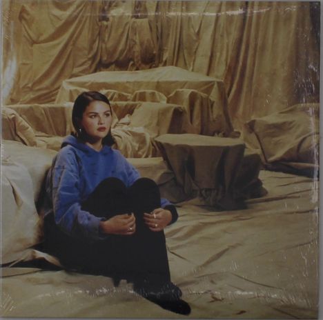 Eine Frau sitzt mit angezogenen Beinen in einem Raum voller mit Tüchern bedeckter Möbel, trägt blauen Hoodie.