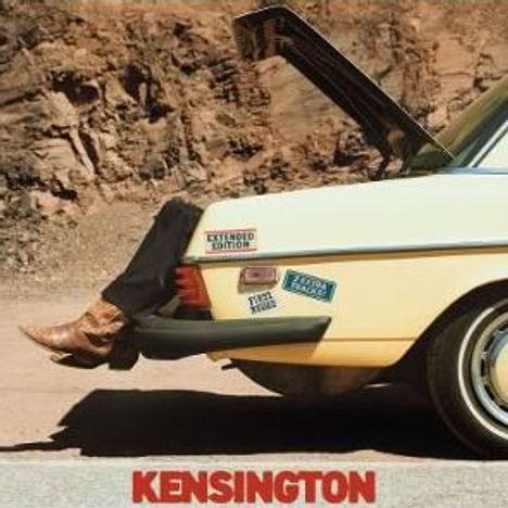 Texte: "Kensington Edition", "Plaza", "Stereo Sound". Beine in Cowboystiefeln ragen aus dem Kofferraum eines gelben Autos.