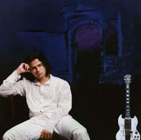 Ein Mann in weißer Kleidung sitzt nachdenklich neben einer weißen E-Gitarre vor dunklem Hintergrund.