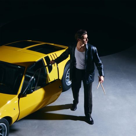 Ein Mann in schwarzer Jacke steht neben einem gelben Auto, hält Trommelstöcke in der Hand und blickt zur Seite.