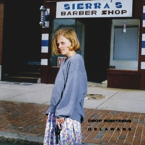 Die Texte „SIERRA'S BARBER SHOP“, „DROP NINETEENS“, „DELAWARE“. Eine junge Frau steht vor dem Friseurladen.