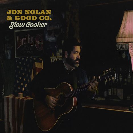 „JON NOLAN & GOOD CO. Slow Cooker”. Ein Mann mit Bart spielt Gitarre in einem dunklen Raum, beleuchtet von einer Lampe.