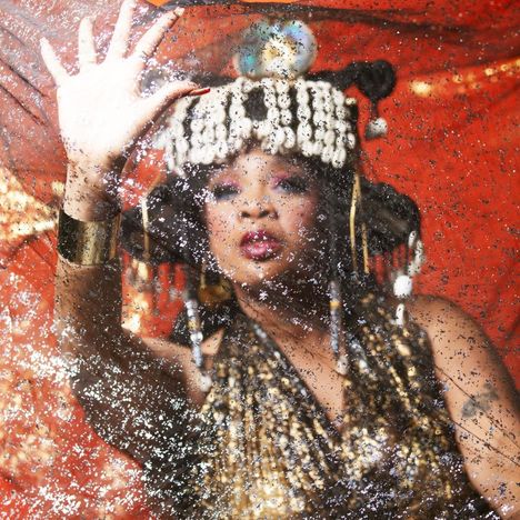 Eine Frau mit opulentem Kopfschmuck und goldenen Accessoires berührt einen glitzernden Schleier vor einem roten Hintergrund.