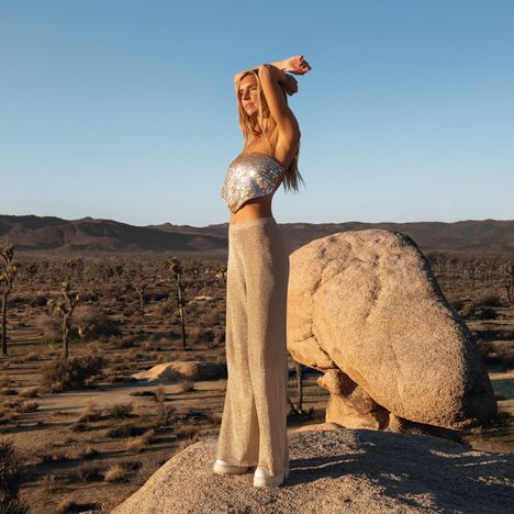 Keine Texte vorhanden. Eine Frau in einem glitzernden Outfit steht auf einem Felsen in der Wüste, im Hintergrund Berge.