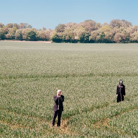 Zwei Personen in einem grünen Feld, eine in dunklem Anzug, die andere in einem Kostüm mit weißer Maske. Wälder im Hintergrund.