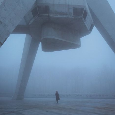 Nebelverhüllte Szene: Eine Person steht auf einem Platz unter monumentaler Betonarchitektur, gefangen in ruhiger Stille.