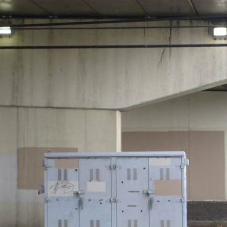 Graffiti auf einem grauen Kasten unter einer Betonbrücke, beleuchtet von zwei Leuchten an der Decke.