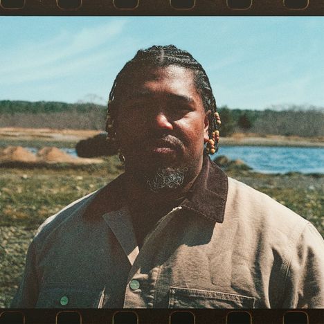 Keine Texte sichtbar. Eine Person mit Jacke und Zöpfen steht draußen, Hintergrund zeigt Wasser und Natur.