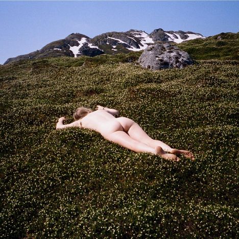 Nackt liegende Person auf blühender Wiese vor schneebedecktem Berg im Hintergrund.