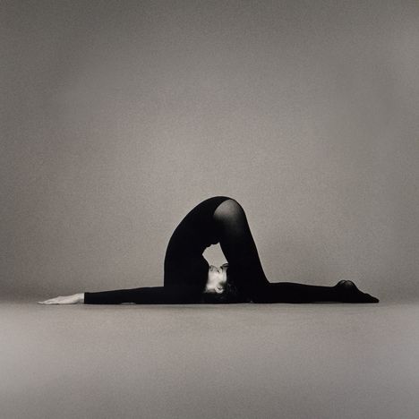 Eine Person in einem schwarzen Ganzkörperanzug macht eine Yoga- oder Ballettpose auf einem grauen Hintergrund.
