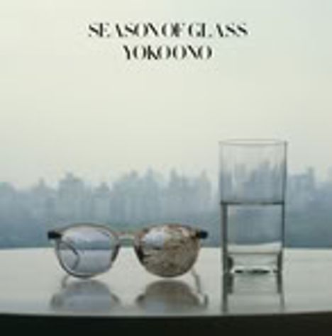 Text: "SEASON OF GLASS YOKO ONO". Eine Brille und ein halbvolles Glas Wasser stehen auf einem Tisch mit Stadt im Hintergrund.