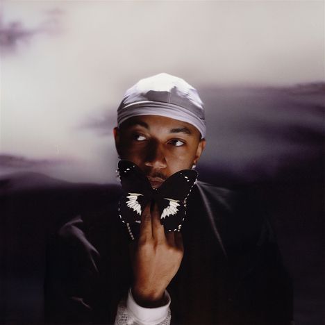 Eine Person mit weißer Mütze hält einen schwarzen Schmetterling vor ihrem Gesicht, der Hintergrund ist verschwommen.