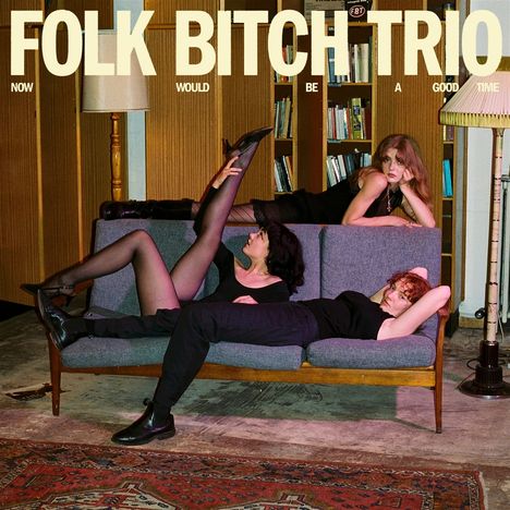 Text: "FOLK BITCH TRIO. NOW WOULD BE A GOOD TIME." Drei Personen posieren in einem Wohnzimmer auf einem Sofa.