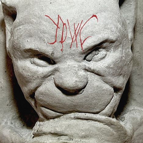 Rote Kritzelei auf der Stirn einer Steinfigur mit grimmigem Gesichtsausdruck und betonten Augenbrauen.