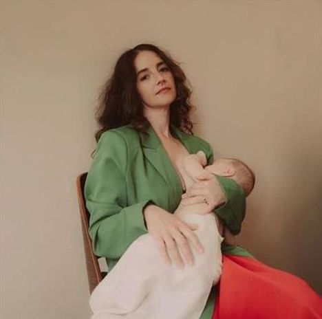 Kein Text vorhanden. Eine Frau in einem grünen Blazer stillt ein Baby, während sie auf einem Stuhl sitzt.