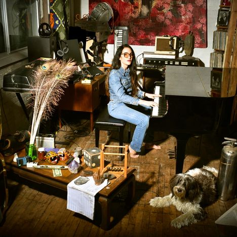 Eine Frau in Jeans spielt Klavier in einem vollen Raum, mit Musikinstrumenten, Dekorationen und einem Hund auf Holzboden.