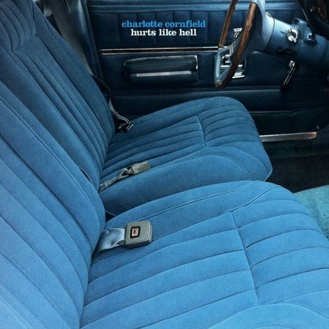 Text: "charlotte cornfield hurts like hell". 
Beschreibung: Innenraum eines Autos mit blauen Stoffpolstern und Lenkrad aus Holz.