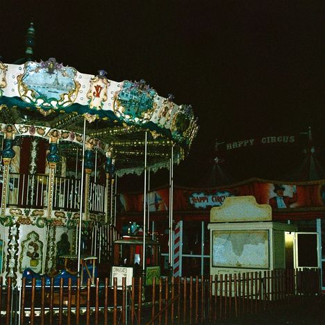 Text: „Happy Circus“. Ein nostalgisches Karussell vor einem Zirkuszelt bei Nacht. Bunte Beleuchtung und Dekor.