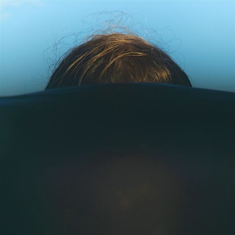 Kein Text. Man sieht den oberen Teil eines Kopfes mit braunem Haar über einer dunklen Oberfläche, vor einem blauen Hintergrund.