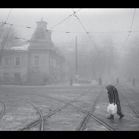 Eine Person mit Tüte überquert schneebedeckte Straßenbahnschienen in nebliger Stadt, die Gebäude wirken alt.