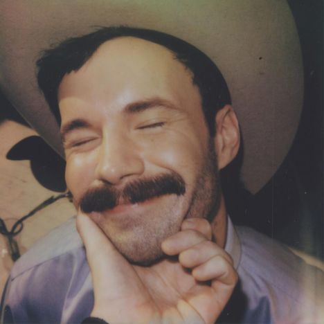 Ein Mann mit Schnurrbart lächelt mit geschlossenen Augen; eine Hand hält sein Kinn.