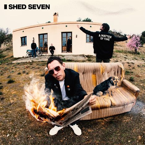 Text: "SHED SEVEN", "A MATTER OF TIME". Ein Mann liest eine brennende Zeitung auf einem Sofa mit einem Hund, vor einem Haus.