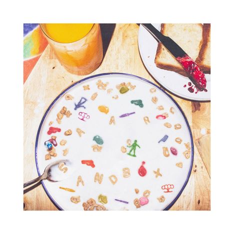 Der Text "SANDBOX" schwimmt in Milch mit bunten Buchstaben. Daneben ein Glas Orangensaft und Toast mit roter Marmelade.