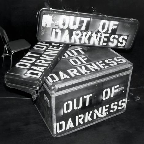 "OUT OF DARKNESS" steht auf mehreren gepackten Reiseboxen in einer Szene mit Stühlen im Hintergrund.