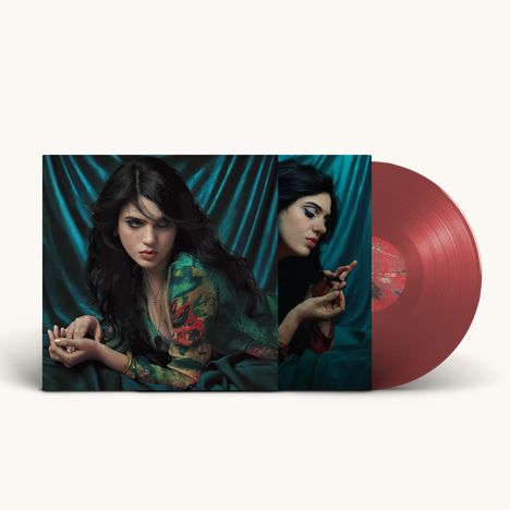 Eine Frau mit dunklem Haar in bunter Kleidung posiert vor blauem Hintergrund. Ein rotes Vinyl ist zu sehen.