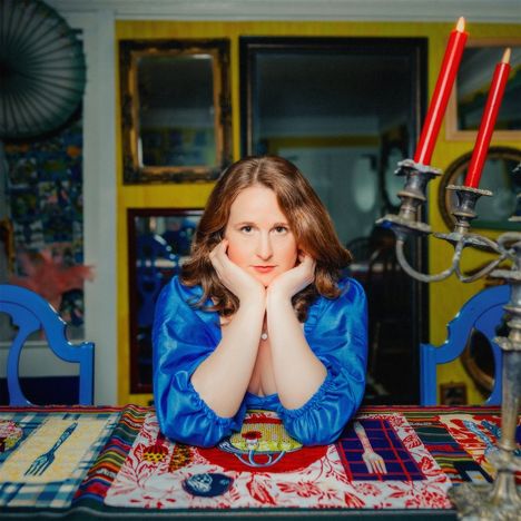 Eine Frau in blauem Kleid stützt ihr Kinn auf ihre Hände am Tisch, bunte Tischdecke, rote Kerzenleuchter links.