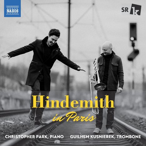 „Hindemith in Paris“ in Gelb. Zwei fröhliche Männer auf Bahngleisen. Einer balanciert, der andere hält eine Posaune.