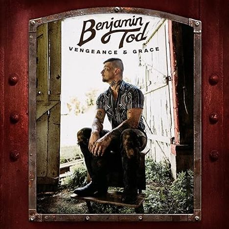 "Benjamin Tod – Vengeance & Grace." Ein Tätowierter sitzt nachdenklich in einem offenen Scheunentor vor ländlicher Kulisse.