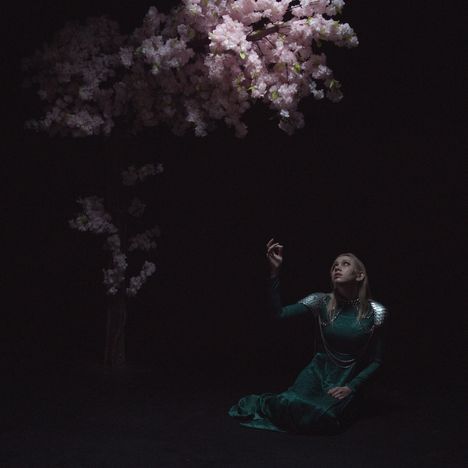 Eine Frau in einem langen, grünen Kleid sitzt im Dunkeln auf dem Boden und schaut zu rosa Blütenzweigen hoch.