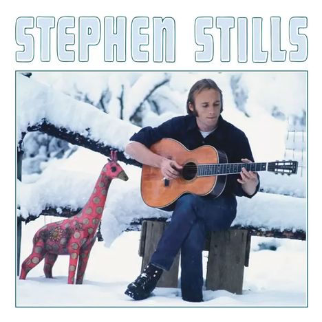 "STEPHEN STILLS" steht oben. Ein Mann spielt Gitarre im Schnee, sitzend auf einer Bank, neben einer bunten Giraffe.
